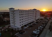 Thumbnail 11 of 13 - A sunset view of a white hotel with a parking lot in front.