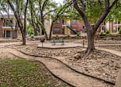 Thumbnail 8 of 35 - A park with a bench and a tree in front of apartment buildings.