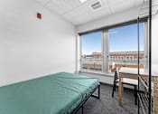 Thumbnail 6 of 61 - A room with a bed, desk, and chair with a window overlooking a cityscape.