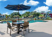 Thumbnail 17 of 38 - A table with chairs and an umbrella is in the foreground of a pool area.