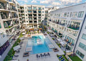 Thumbnail 25 of 27 - A large swimming pool is surrounded by lounge chairs and umbrellas in a courtyard surrounded by apartment buildings.