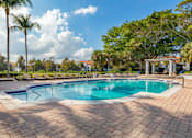 Thumbnail 9 of 28 - A large outdoor swimming pool surrounded by a brick patio and palm trees.