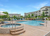 Thumbnail 20 of 29 - A large outdoor pool with a hot tub and a building in the background.