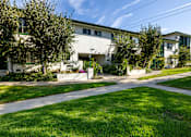 Thumbnail 4 of 12 - A white building with green trim and a sign that says "The Oaks" is surrounded by trees and grass.