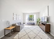 Thumbnail 5 of 30 - A living room with a grey couch, a wooden table, a rug, and a window with a view of the outside.