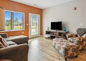 Thumbnail 5 of 27 - A living room with a grey couch, a brown patterned ottoman, and a flat screen TV.