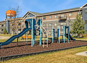 Thumbnail 21 of 27 - A playground with a blue slide and a brown bench in front of a building.