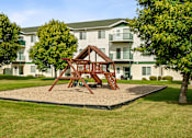 Thumbnail 21 of 29 - A playground with a swing set in the middle of a grassy area in front of apartment buildings.