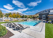 Thumbnail 94 of 136 - a swimming pool with patio furniture and a building with mountains in the background at The Flats at Riverwoods, Provo, UT