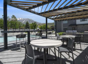 Thumbnail 102 of 136 - a patio with tables and chairs with a pool and mountains in the background at The Flats at Riverwoods, Provo, Utah