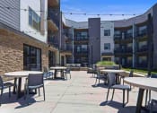 Thumbnail 11 of 136 - a patio with tables and chairs in front of an apartment building at The Flats at Riverwoods, Provo, 84604