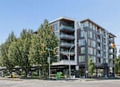 Thumbnail 27 of 28 - A modern multi-story building with balconies is situated on a street corner with a traffic light.