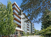 Thumbnail 28 of 28 - A modern multi-story building with balconies is surrounded by trees and greenery.