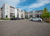Thumbnail 6 of 21 - A parking lot in front of a multi-story apartment building with cars parked.