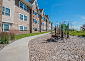 Thumbnail 5 of 8 - A playground with a swing set is located in front of a building with a paved walkway.