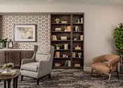Thumbnail 17 of 19 - A living room with a grey chair, a brown chair, a bookshelf, and a potted plant.