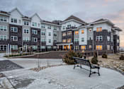 Thumbnail 10 of 11 - A bench sits in front of a building with a mix of white and grey balconies.