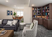 Thumbnail 13 of 24 - A living room with a grey sofa, a grey armchair, a coffee table, and a bookshelf filled with books.