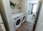 Thumbnail 12 of 29 - A laundry room with two washing machines and a shelf with towels and plants.