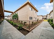 Thumbnail 7 of 15 - A concrete pathway leads to a building with a metal fence and a small tree in front.