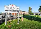 Thumbnail 1 of 14 - A sign for Villa De La Rosa Apartment Homes stands in front of a building.
