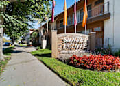 Thumbnail 3 of 70 - Sunset Polinte Apartment Homes sign with flags and red bushes.