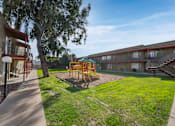 Thumbnail 4 of 18 - A playground with a slide and swings is surrounded by grass and trees.