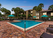 Thumbnail 14 of 23 - A pool surrounded by a brick patio and palm trees at Boca Colony Apartments, Boca Raton, FL