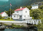 Thumbnail 12 of 17 - A white house with a red roof is situated by a body of water with a bridge leading to it.