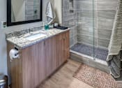 Thumbnail 41 of 43 - Apartments For Rent In Downtown Kalamazoo, MI - Large Bathroom With Grey Tile Shower And Glass Doors, Granite Countertops, Large Vanity Mirror, And Wood-Inspired Cabinets And Flooring