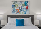 Thumbnail 3 of 30 - a bedroom with a bed with a blue pillow and a colorful painting above it at Tivoli Green Apartments & Townhomes, Mount Pleasant, WI