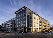 Thumbnail 1 of 31 - A modern apartment building with a mix of dark and light facades is situated on a street corner with cars parked along the curb.