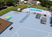Thumbnail 23 of 27 - A basketball court with a blue surface and white lines, surrounded by a fence and with a pool in the background.