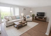 Thumbnail 6 of 10 - A living room with a white couch, a white chair, a wooden coffee table, and a television.