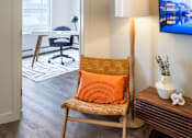 Thumbnail 13 of 30 - A chair with an orange pillow is in the foreground of a room with a television on the wall.
