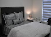 Thumbnail 1 of 9 - A bed with a grey comforter and pillows is in a room with a window and a lamp at The Laurent at Laurel Apartments, Edmonton, AB, T6T 2T5