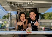 Thumbnail 4 of 10 - A man and woman are standing behind a food truck counter, smiling and pointing at the camera.