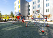 Thumbnail 6 of 38 - A playground with a red and grey climbing structure in front of a building.