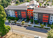 Thumbnail 2 of 21 - A modern building with solar panels on the roof is surrounded by trees and a street with cars.