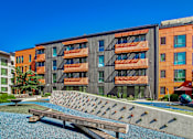 Thumbnail 19 of 21 - A modern building with orange and grey balconies is surrounded by a small pool and a wooden bench.