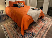 Thumbnail 2 of 18 - A bedroom with a bed covered in an orange bedspread and a rug on the floor.