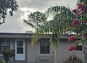 Thumbnail 3 of 33 - A house with a white door and windows is surrounded by greenery and a paved walkway.