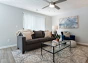Thumbnail 6 of 35 - A living room with a grey couch, a glass coffee table, and a ceiling fan.