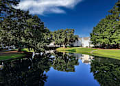 Thumbnail 35 of 42 - A serene landscape with a pond, trees, and a white building in the distance.