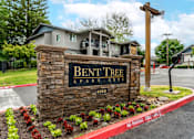 Thumbnail 1 of 31 - Entrance to Bent Tree Apartments with stone monument sign and large trees providing a cozy atmosphere