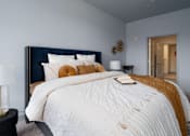 Thumbnail 28 of 112 - A bed with a white comforter and a blue headboard with a gold pillow and a book on it. at The Residences at Tesson Ridge Apartments, Missouri