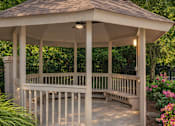 Thumbnail 17 of 23 - A gazebo with a brown roof and white pillars is surrounded by greenery and flowers.