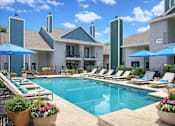 Thumbnail 19 of 25 - Dog-Friendly Apartments in Churchill Estates, TX - Apex Apartments - Swimming Pool With Lounge Chairs, Blue Umbrellas, Colorful Planters, And Surrounding Residential Buildings.