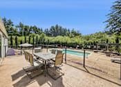 Thumbnail 8 of 40 - A table and chairs are set up on a patio with a pool and trees in the background.