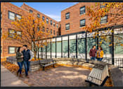 Thumbnail 4 of 20 - A couple is standing on a brick patio in front of a brick building with a tree with orange leaves.
